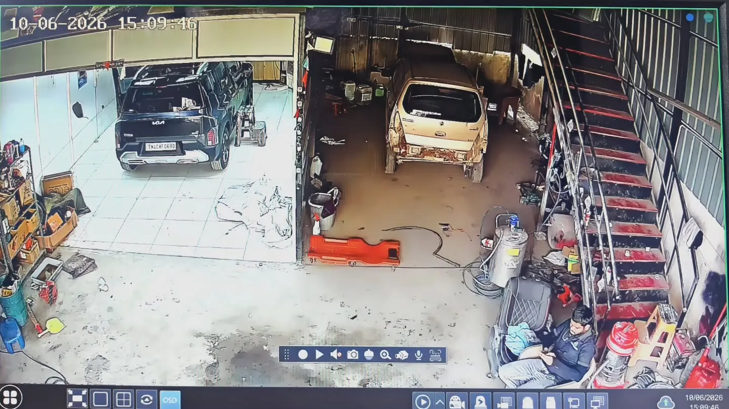 High-angle wide security camera view from a Gopix 5MP IP camera capturing the main automotive service bays inside The Detailing Guru in Thandalam, showing a black Kia and a silver Maruti Suzuki parked on the service floors with active detailing tools and machinery nearby.