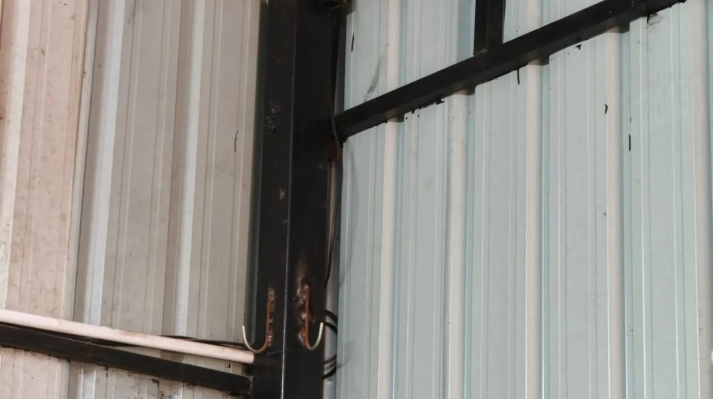 High-angle indoor view of a white Gopix bullet CCTV camera neatly mounted on a surface junction box in a high corner under a corrugated metal roof structure at an automotive garage workshop.