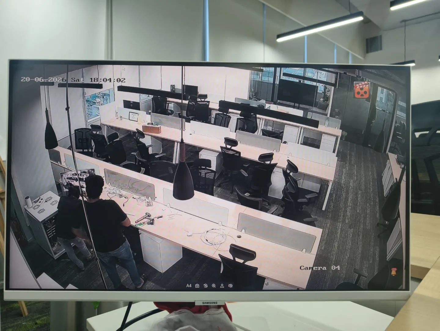 A detailed single-channel full-screen monitor view showing real-time, high-resolution indoor office surveillance of long tech workbenches and seating clusters.