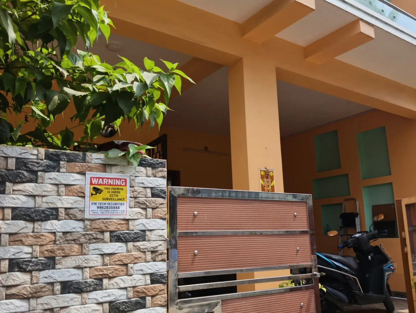 Eye Tech Securities CCTV warning sticker pasted on a residential building's stone-clad compound wall next to the main entrance gate.