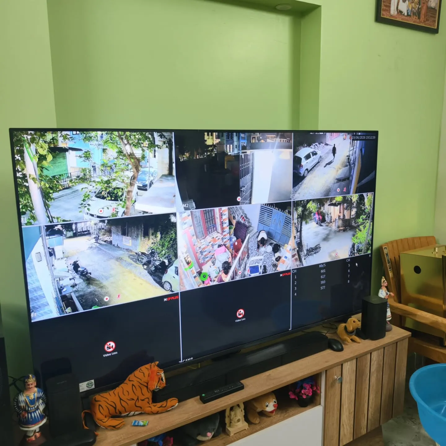 A large living room TV display showing a 6-channel live matrix view of a newly installed Cpplus 2MP HD Dual Light security camera system covering the home's outdoor street lanes, entrance gate, and compound areas.