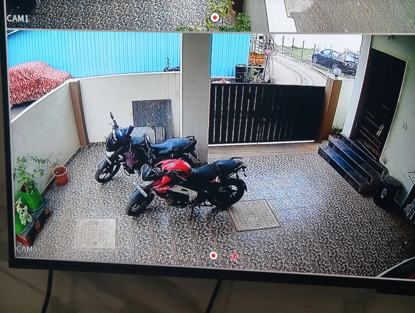 A sharp, high-definition live feed from a CP Plus 5MP camera capturing parked two-wheelers, the main entrance gate, and the porch access steps.