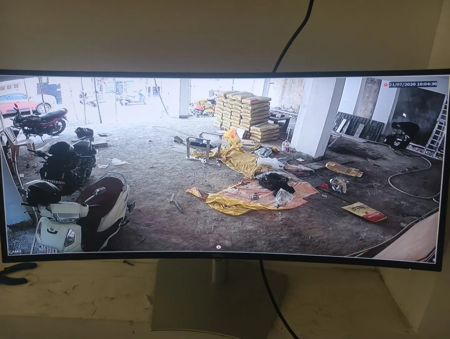 A wide-angle monitor feed from a CP Plus Dual Light camera covering the covered parking bay, vehicle storage, and construction materials at a residential site.