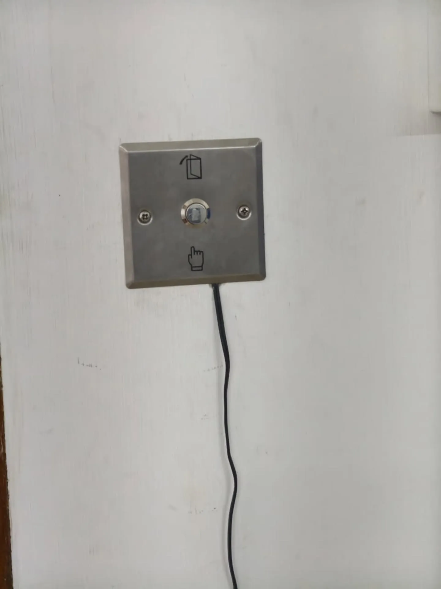 ​A sleek, stainless steel push-to-exit switch cleanly mounted on an interior office wall with a black connection cable running down.