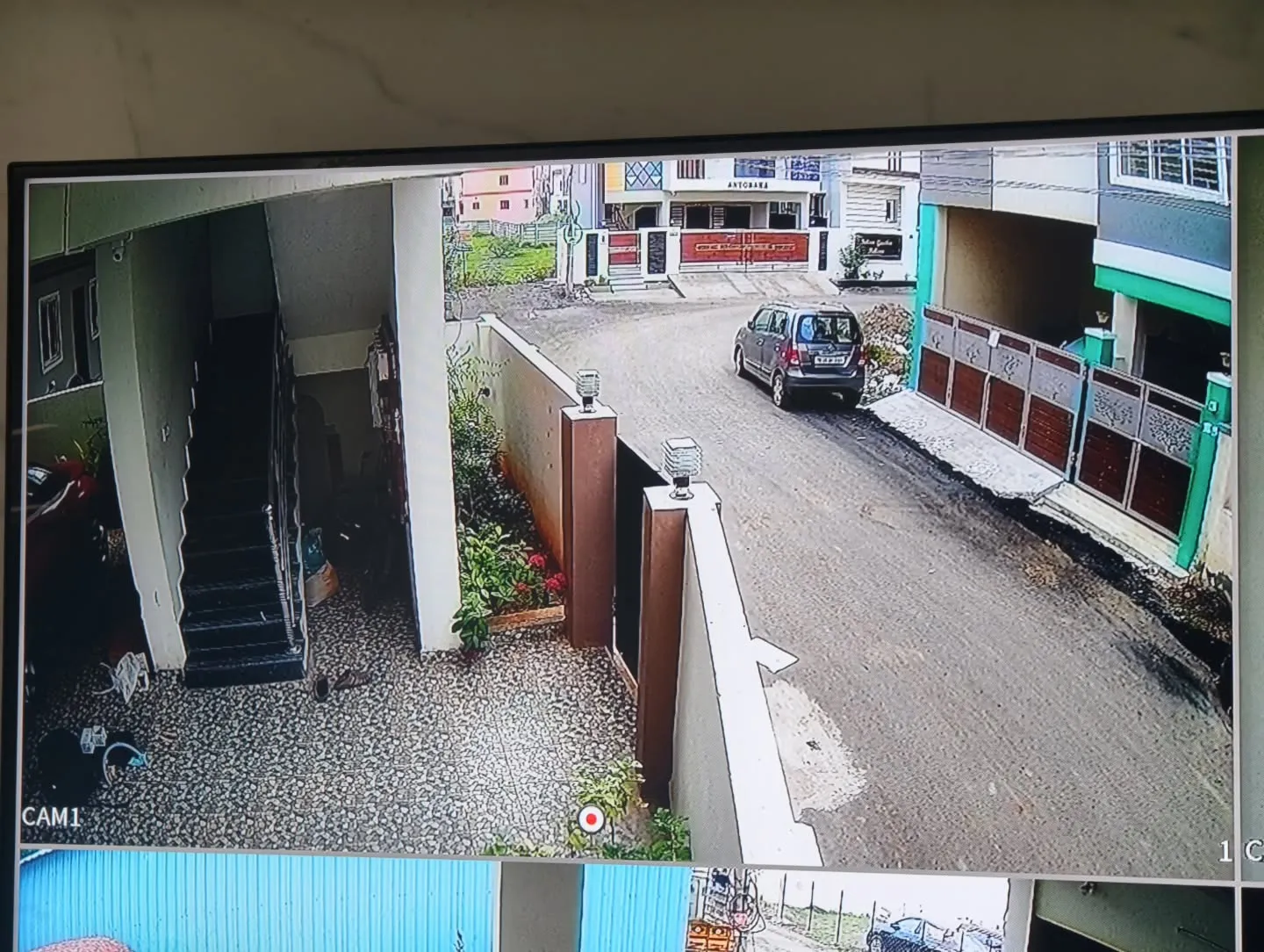 A wide-angle live video display from a CP Plus 5MP outdoor camera monitoring the residential main gate, compound wall, and front street approach.