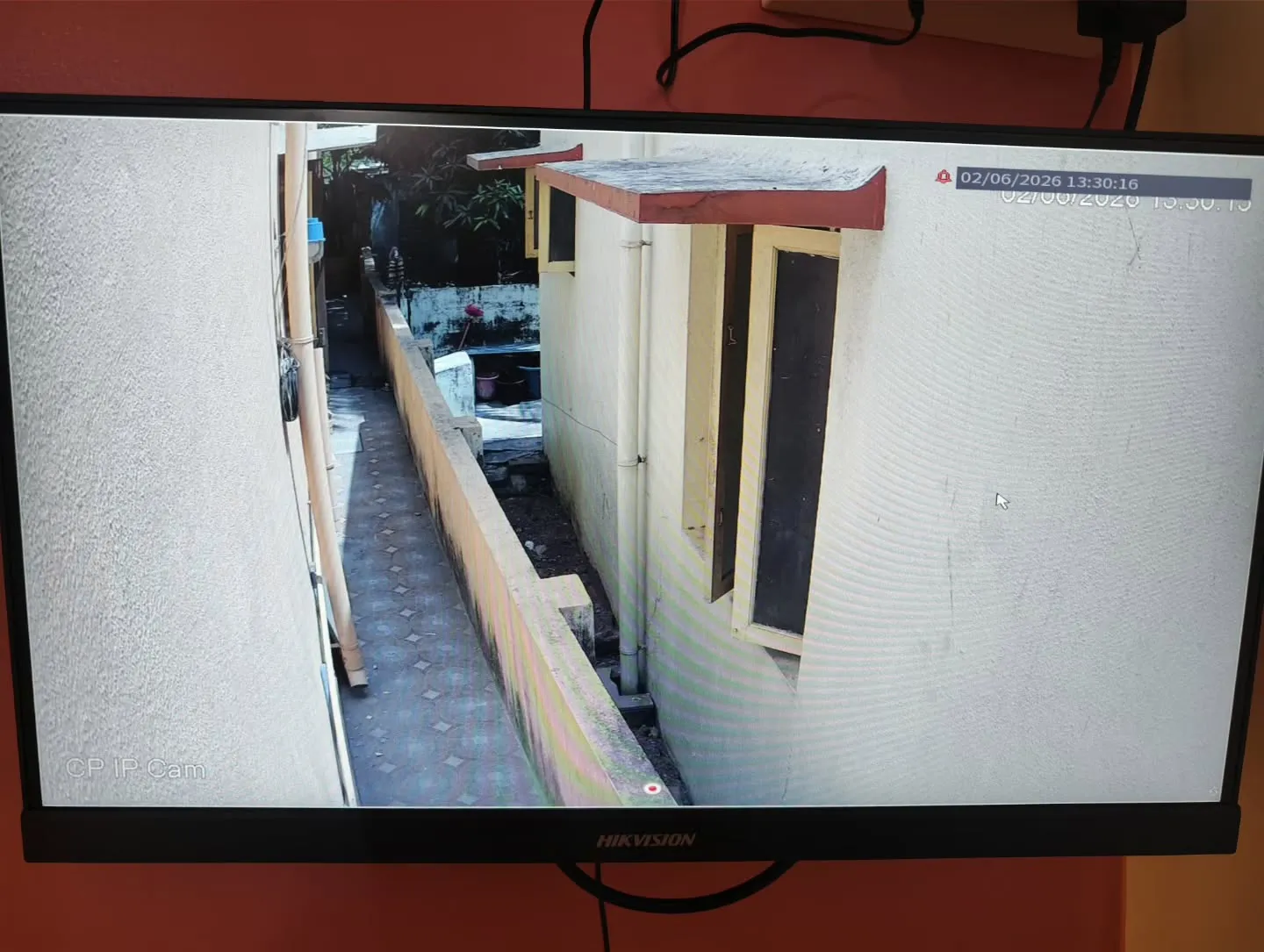 CP Plus 4MP STQC IP camera view of a narrow residential side setback, showing the building wall, windows, and compound boundary.