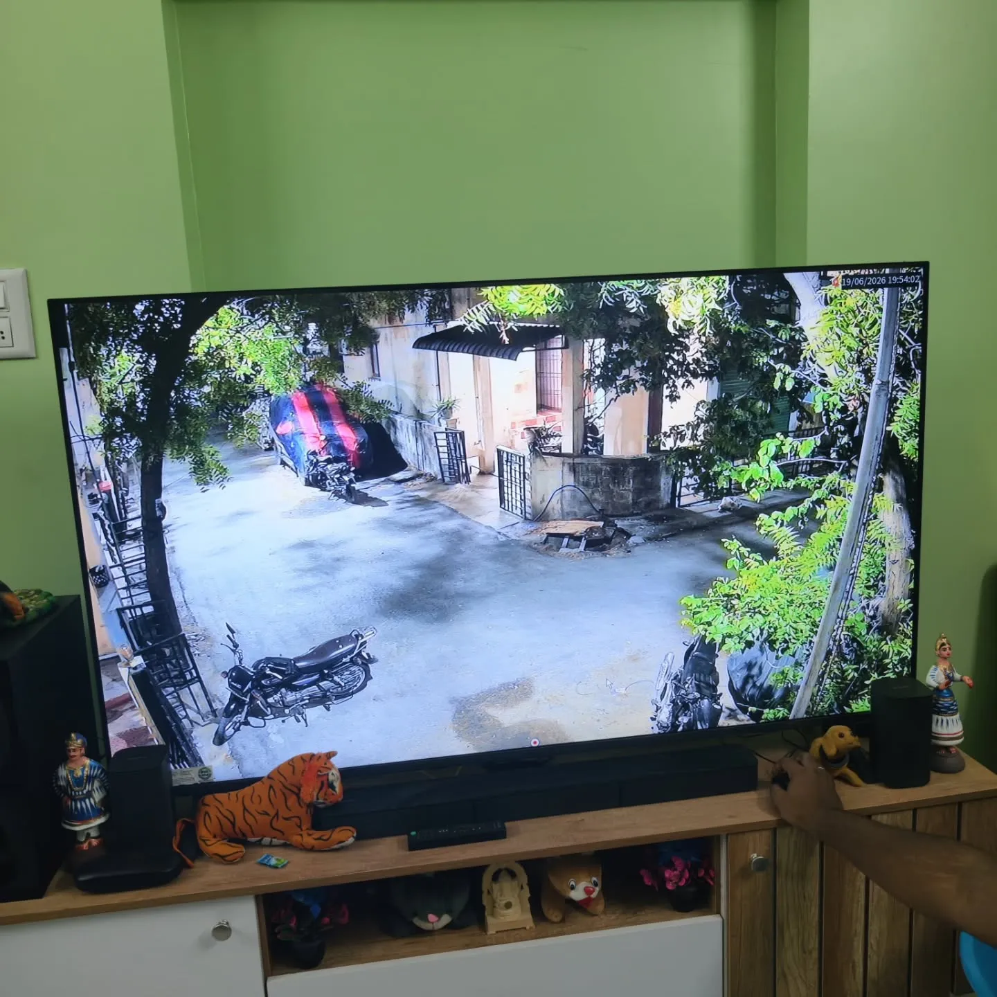 ​A full-screen single-channel live feed on a home smart TV, showing clear outdoor surveillance coverage from a Cpplus 2MP HD Dual Light camera monitoring a residential street lane, opposite houses, and a parked motorcycle.