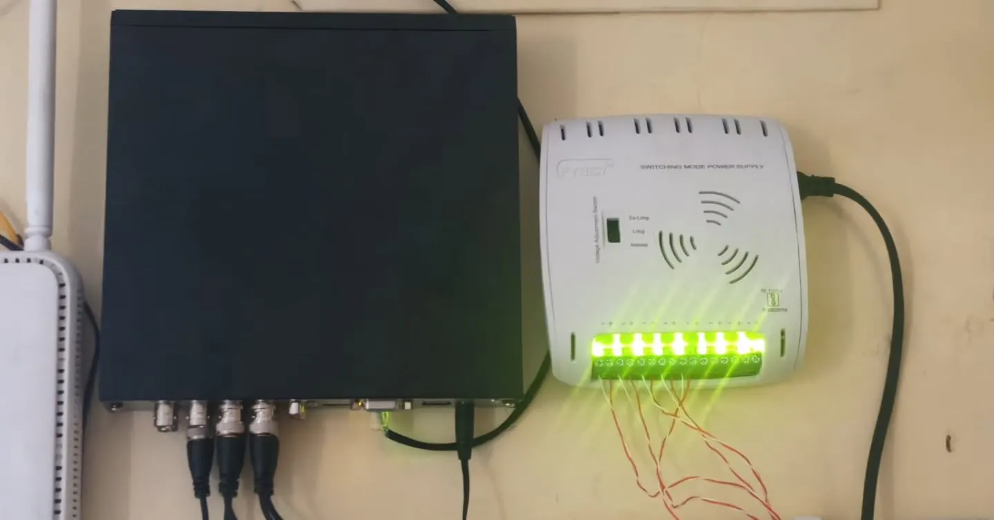 Wall-mounted CCTV backend setup featuring a black 4-channel DVR with connected video baluns, a white multi-channel switching mode power supply (SMPS) with active green LED status lights, and a Wi-Fi router fixed directly to the wall.