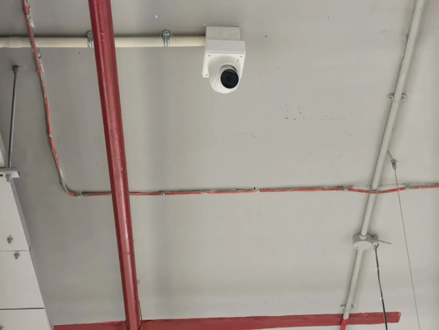 ​A ceiling-mounted Hikvision dome CCTV camera anchored beside fire sprinkler pipes with industrial-style exposed infrastructure.