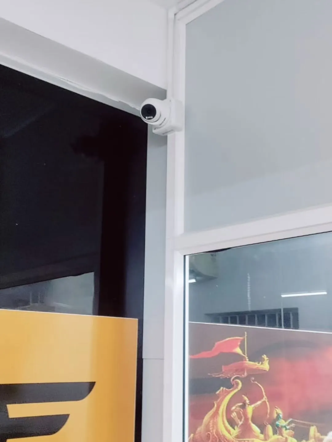 ​A compact white Hikvision dome security camera neatly installed at the intersection of a wall and ceiling partition structure.