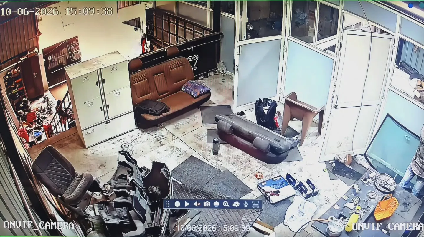 High-angle wide indoor security camera view from a Gopix 5MP IP camera capturing the main reception area, seating lobby, and workspace floor inside The Detailing Guru garage in Thandalam, featuring car detailing seats, structural partitions, and premium equipment on the floor.