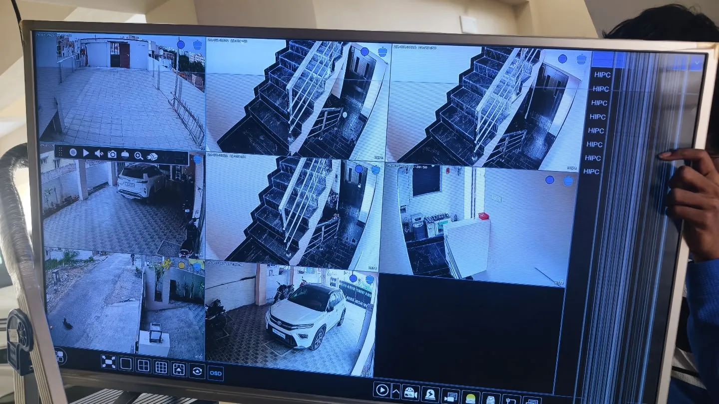 ​A multi-channel CCTV monitor display running live feeds of the parking lot, staircases, and entrance at Thendral Avenue Apartment.