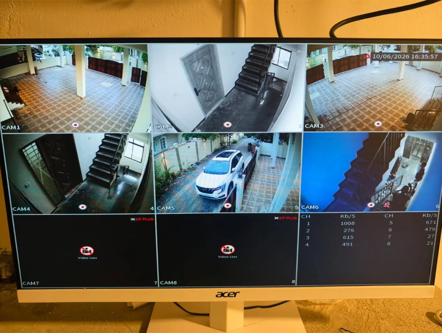 An Acer monitor displaying a 6-channel live security feed from CP Plus 5MP cameras monitoring apartment parking areas, entrance gates, and stairwells.