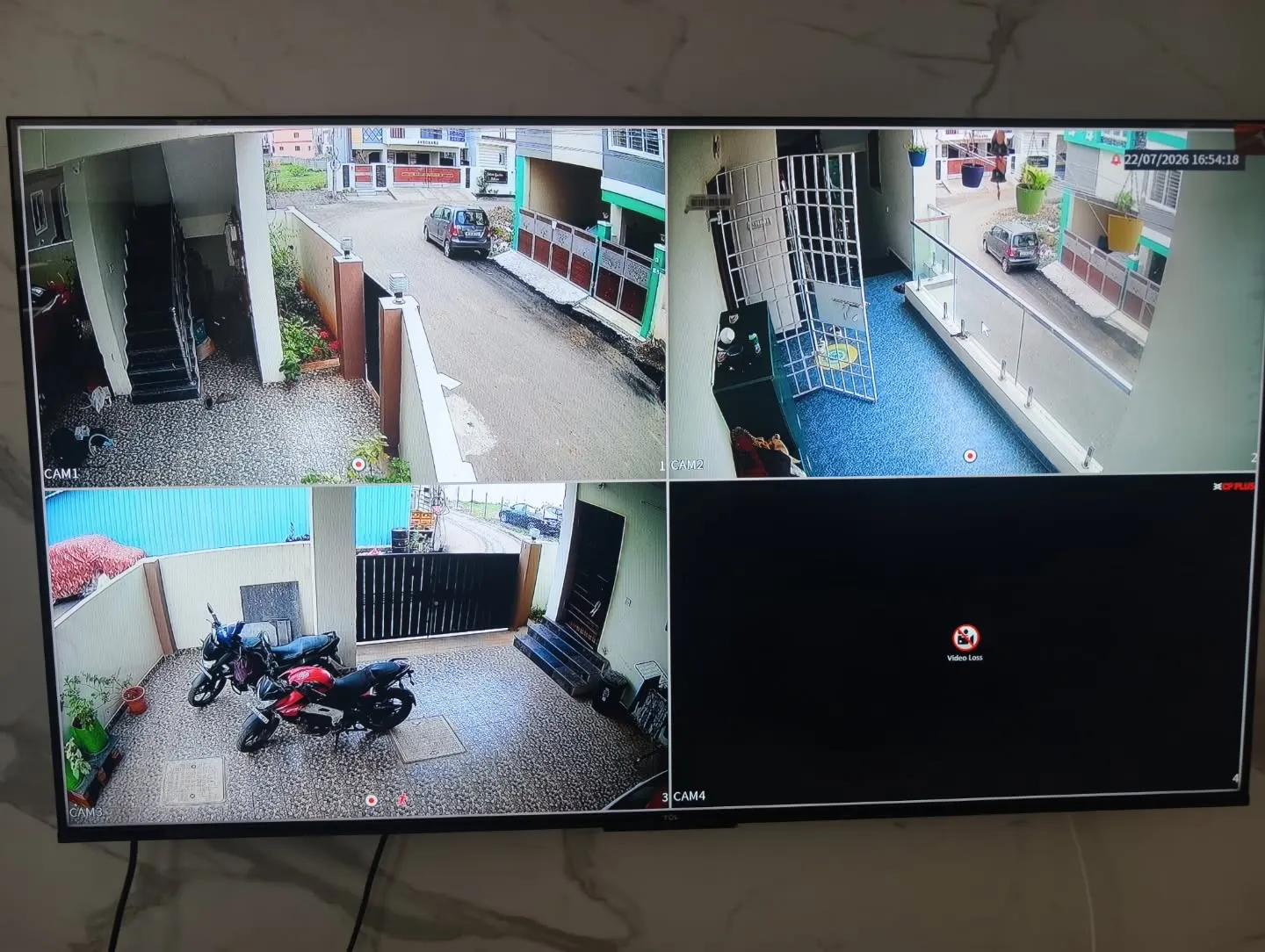 ​A 4-channel monitor display showing a split-screen live view from CP Plus 5MP Dual Light cameras installed across the entrance gate, balcony, and ground-floor parking area