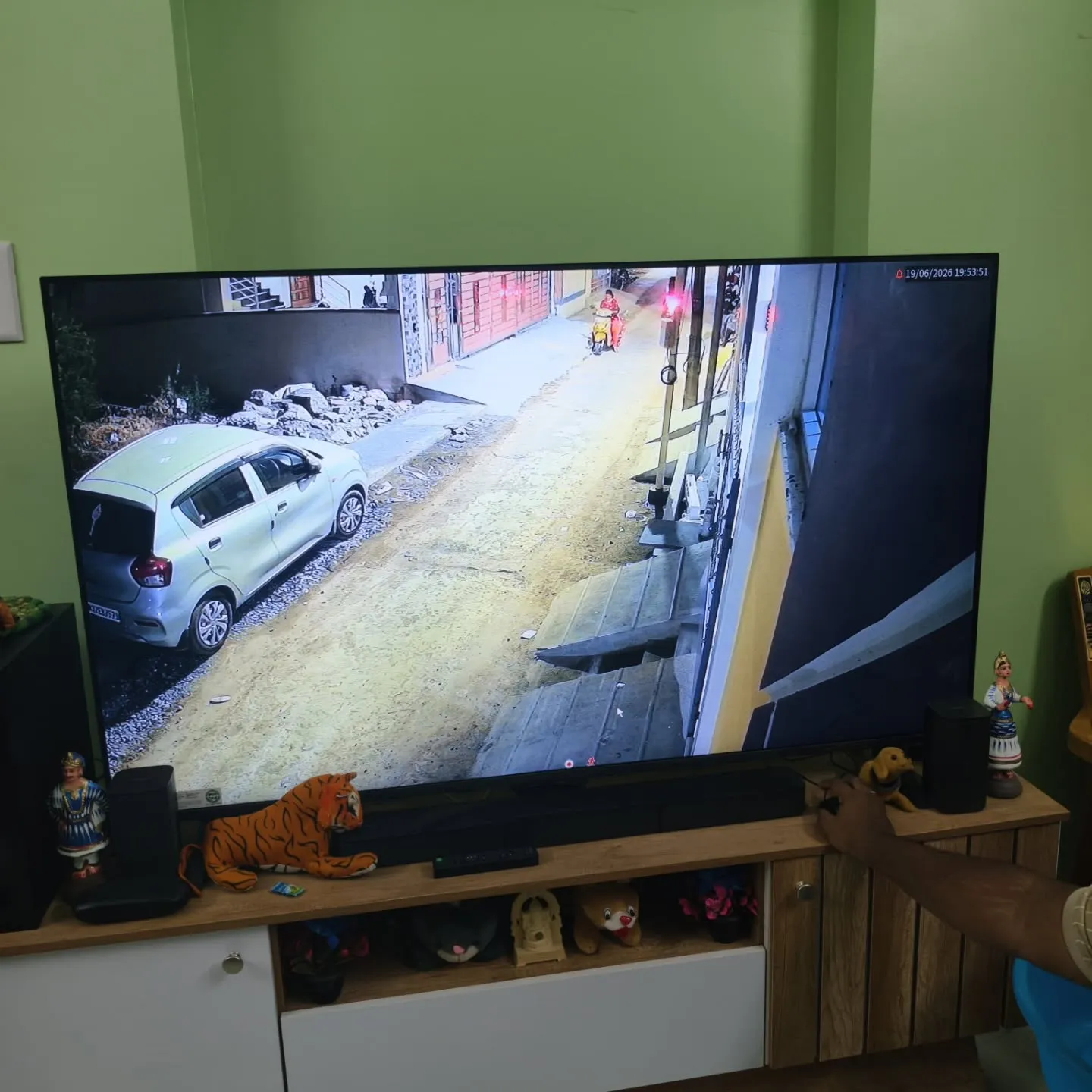 TV screen showing a Cpplus 2MP HD Dual Light camera feed monitoring a street lane entrance, house ramp, and parked hatchback car.